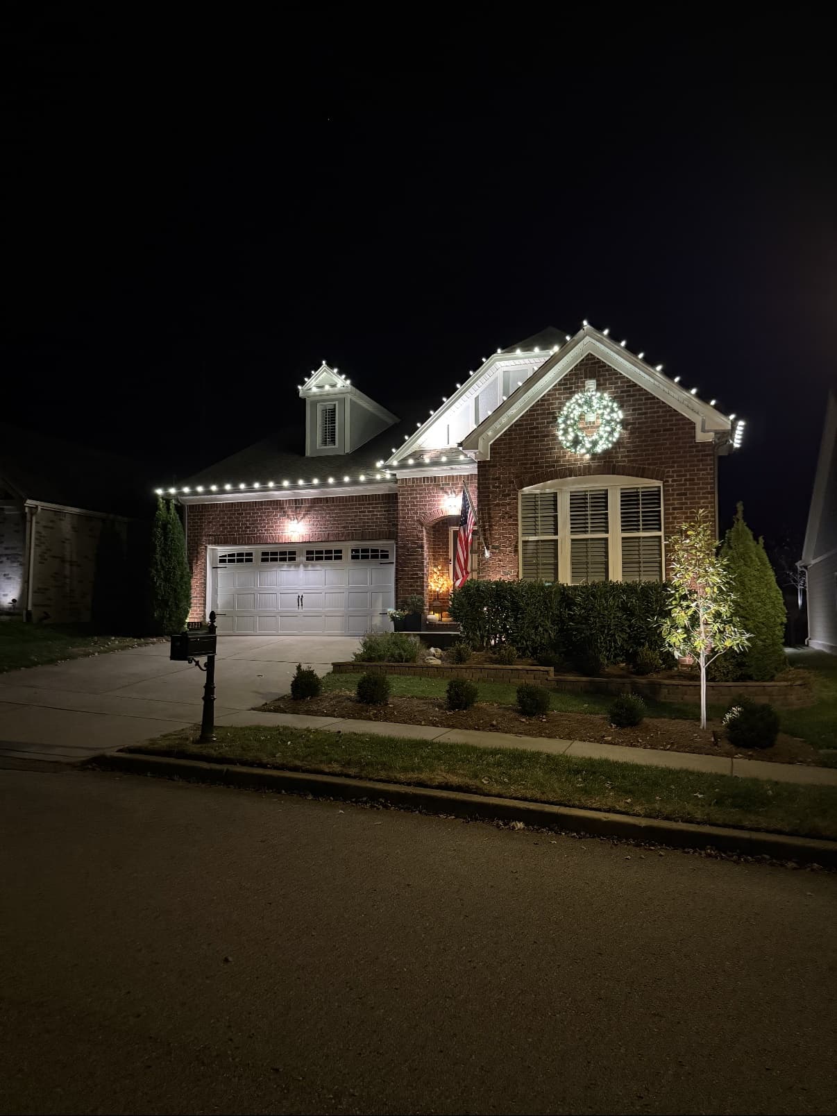 Two-story brick craftsman home with warm white C9 rooflines and a lit wreath at dusk